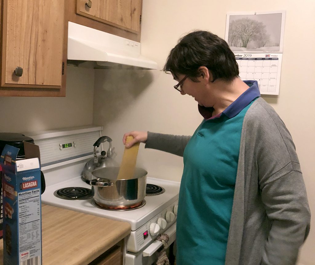 A woman in a blue shirt and gray cardigan with short dark hair and glasses places pasta into a pot of boiling water.